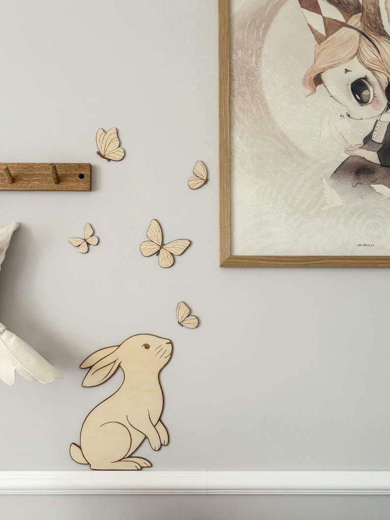 May include: Wooden wall decor featuring a light brown rabbit looking up at a group of wooden butterflies. A framed illustration with a child-like figure is on the wall. A wooden coat rack is also visible.