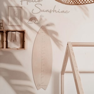 May include: A light wood surfboard-shaped growth chart with the words "Ocean Child" and "Oliver" on it. The chart is hanging on a white wall with a wooden bed frame and a woven light fixture in the background. The wall also has a rainbow and clouds, the words "Hello Sunshine", and a framed picture of a baby.