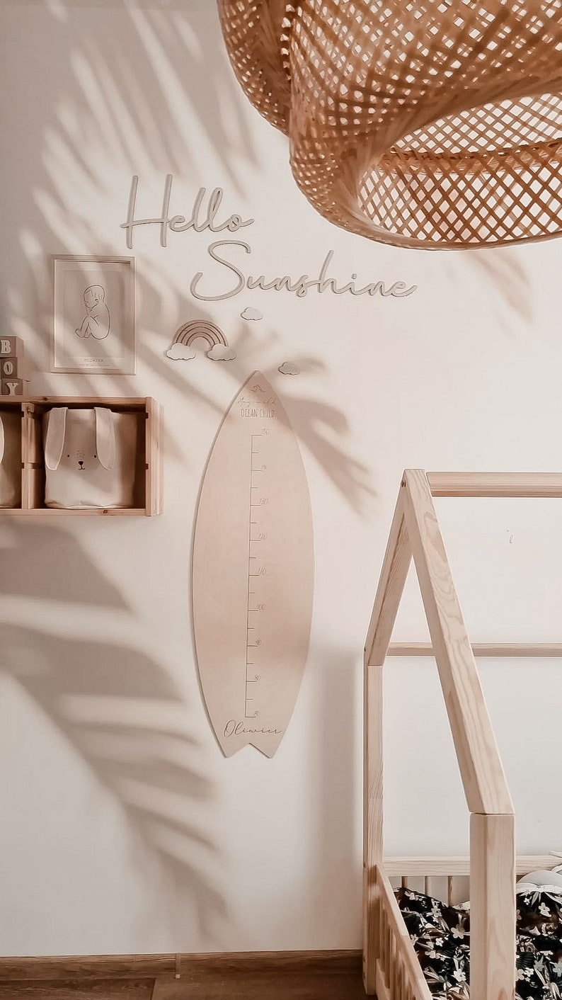 May include: A light wood surfboard-shaped growth chart with the words "Ocean Child" and "Oliver" on it. The chart is hanging on a white wall with a wooden bed frame and a woven light fixture in the background. The wall also has a rainbow and clouds, the words "Hello Sunshine", and a framed picture of a baby.