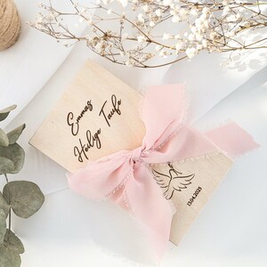 May include: A wooden box with a pink ribbon tied around it. The box is engraved with the text "Emma's Holy Baptism" and the date "13.04.2023".