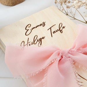 May include: A wooden box with a pink ribbon tied around it. The box is engraved with the text "Emma's Heilige Taufe".