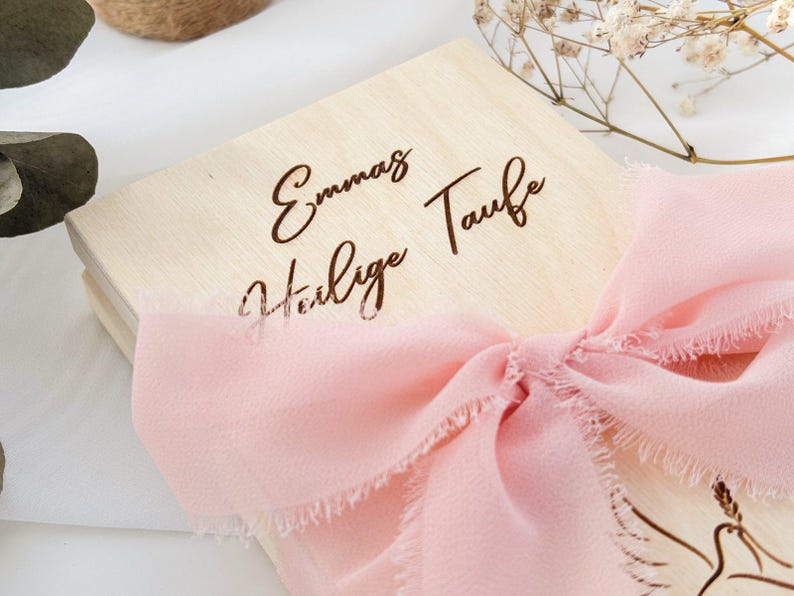 May include: A wooden box with a pink ribbon tied around it. The box is engraved with the text "Emma's Heilige Taufe".