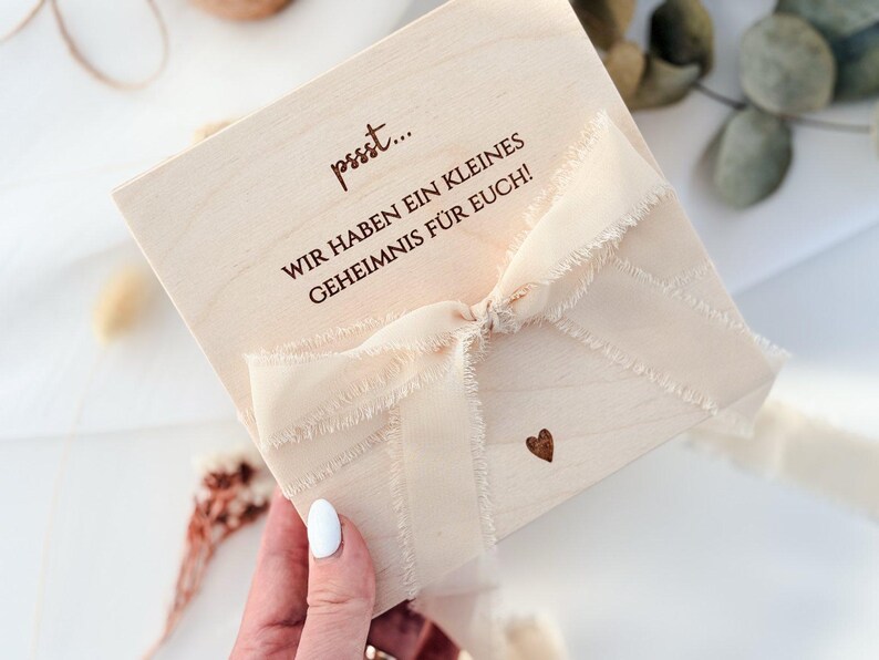 May include: A light-colored wooden box tied with a cream-colored ribbon. The box has text in brown ink that reads, "pssst... WIR HABEN EIN KLEINES GEHEIMNIS F&Uuml;R EUCH!" A small brown heart is printed on the box.