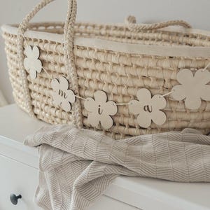 May include: A woven basket with a braided handle, decorated with a flower garland spelling out a name. A folded beige blanket rests on a white dresser. The basket is a light beige color.