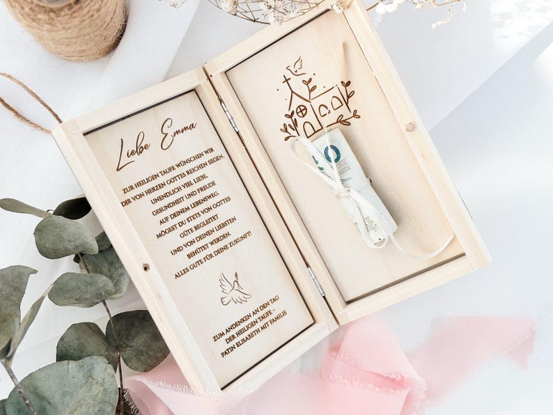 May include: A wooden box with a hinged lid, engraved with a church and a dove. The box is open, revealing a white envelope with a ribbon tied around it. The text on the box reads "Liebe Emma" and includes a message about a baptism.