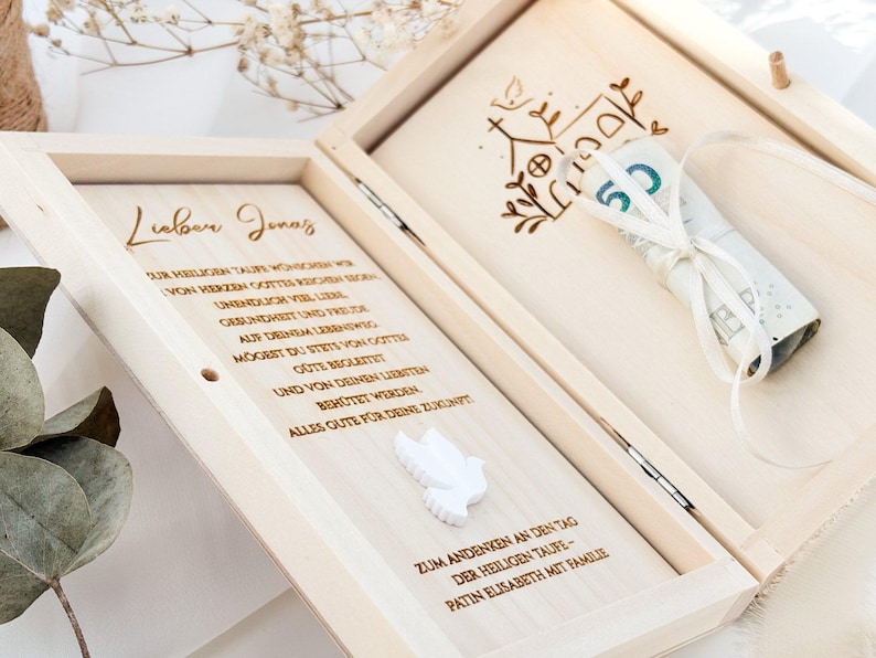 Pu&ograve; includere: Una scatola di legno con un coperchio incernierato, incisa con un messaggio e una colomba. La scatola contiene una banconota arrotolata legata con un nastro bianco. Il testo sulla scatola recita "Lieber Jonas" e "Zum Andenken an den Tag der Heiligen Taufe - Patin Elisabeth mit Familie".
