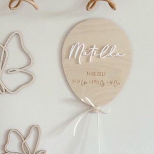 May include: A wooden balloon-shaped sign with the name "Matilda" in white script. The sign includes the date 24.02.2023 and measurements. A white ribbon is tied at the bottom. Two wooden butterfly cutouts are on the left.