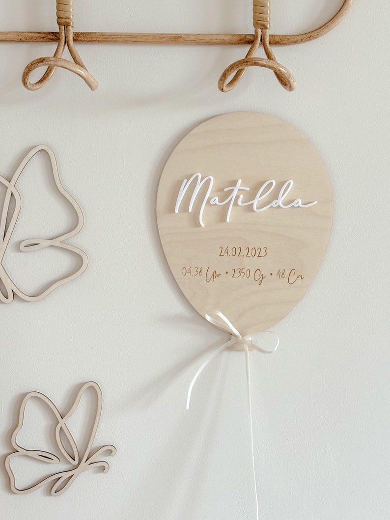 May include: A wooden balloon-shaped sign with the name "Matilda" in white script. The sign includes the date 24.02.2023 and measurements. A white ribbon is tied at the bottom. Two wooden butterfly cutouts are on the left.