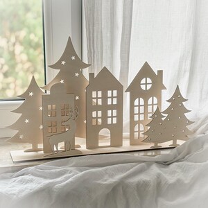 Puede incluir: Una escena de pueblo navide&ntilde;o de madera con casas, &aacute;rboles y un reno recortados. Las casas tienen recortes de ventanas y los &aacute;rboles tienen recortes en forma de estrella. La escena se muestra sobre una base de madera, ideal para la decoraci&oacute;n navide&ntilde;a.