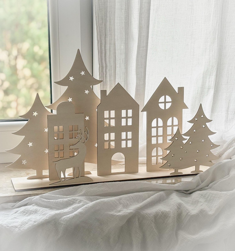 Puede incluir: Una escena de pueblo navide&ntilde;o de madera con casas, &aacute;rboles y un reno recortados. Las casas tienen recortes de ventanas y los &aacute;rboles tienen recortes en forma de estrella. La escena se muestra sobre una base de madera, ideal para la decoraci&oacute;n navide&ntilde;a.