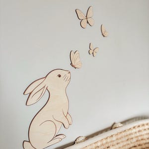 May include: Wooden wall art featuring a bunny and butterflies. The bunny is light brown with a darker outline, looking upwards towards several wooden butterflies. The butterflies are in various sizes and are also light brown with darker outlines. The art is mounted on a light gray wall.