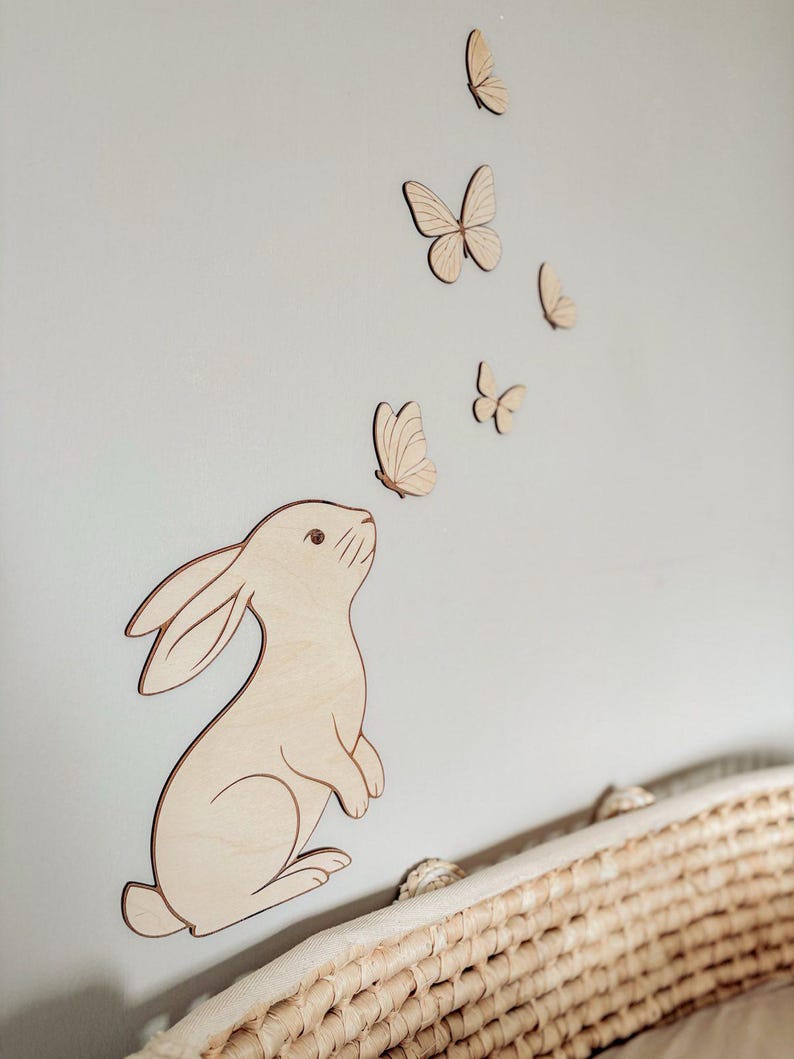 May include: Wooden wall art featuring a bunny and butterflies. The bunny is light brown with a darker outline, looking upwards towards several wooden butterflies. The butterflies are in various sizes and are also light brown with darker outlines. The art is mounted on a light gray wall.