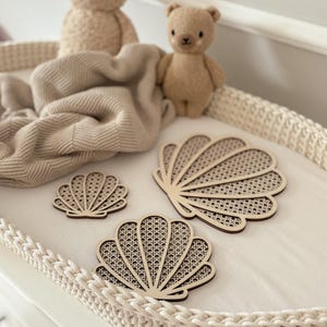 May include: Three wooden seashell-shaped coasters with a woven cane design are displayed on a white surface. Two small teddy bears and a beige blanket are in the background. The coasters vary in size and have a light brown color.