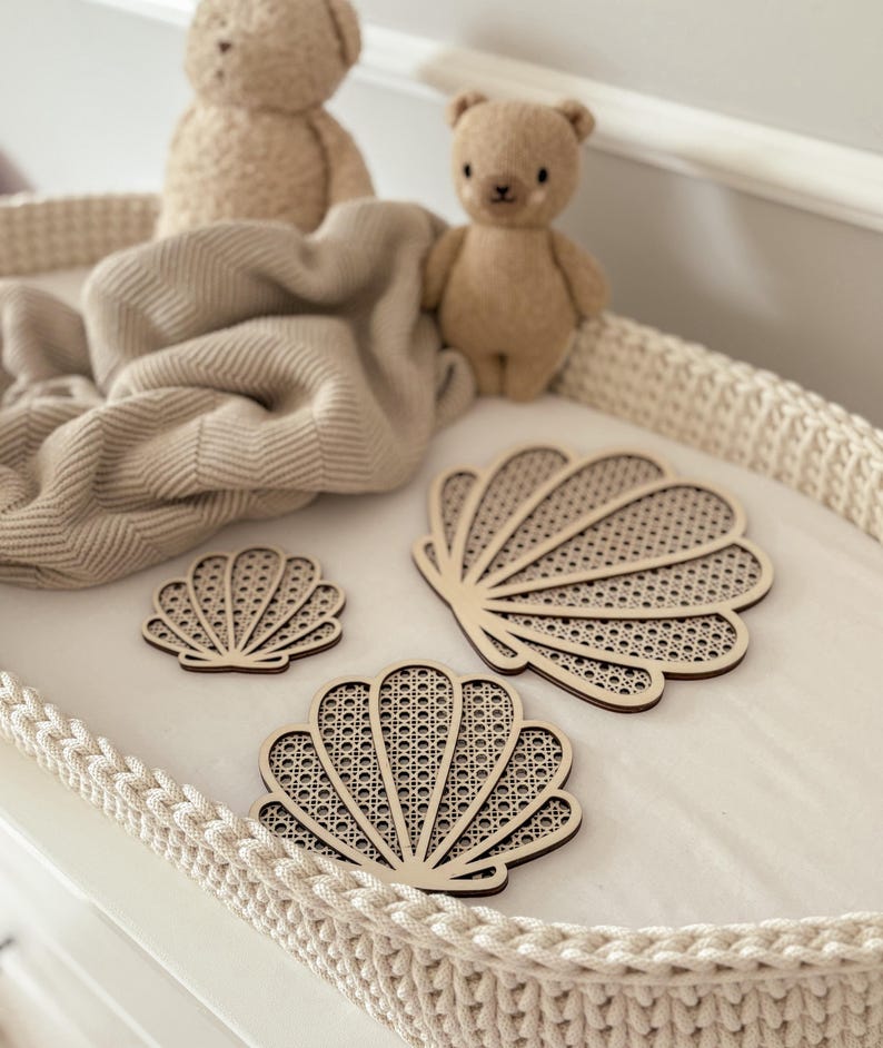May include: Three wooden seashell-shaped coasters with a woven cane design are displayed on a white surface. Two small teddy bears and a beige blanket are in the background. The coasters vary in size and have a light brown color.
