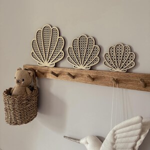 May include: Three wooden seashell wall decorations with a woven cane design are mounted above a wooden coat rack. A small woven basket with a teddy bear hangs on the left. A white bird decoration is on the right.