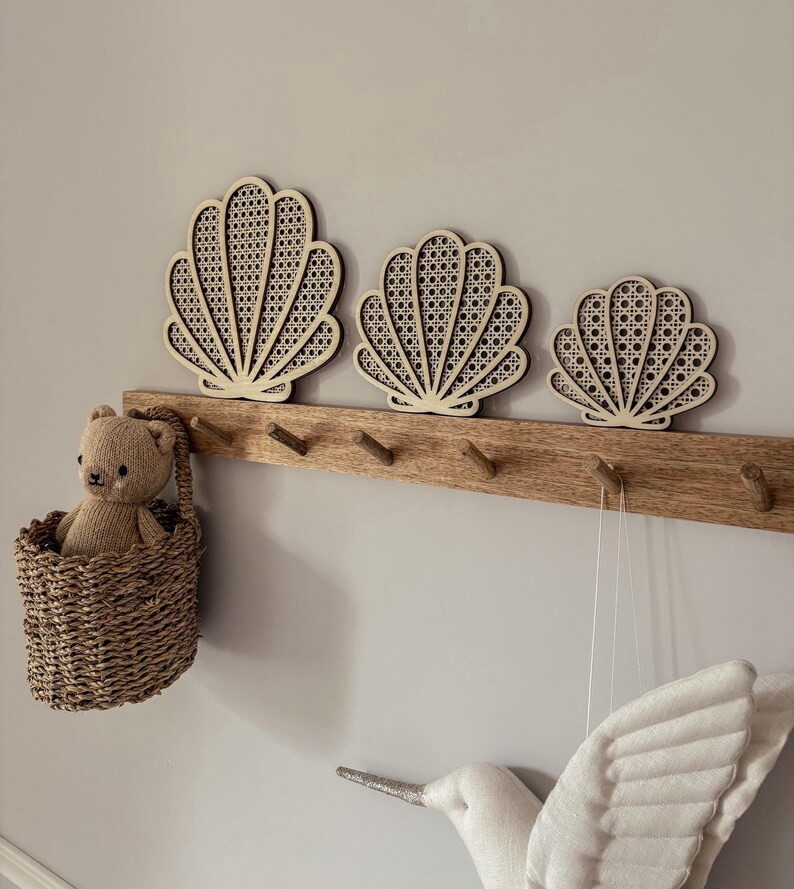 May include: Three wooden seashell wall decorations with a woven cane design are mounted above a wooden coat rack. A small woven basket with a teddy bear hangs on the left. A white bird decoration is on the right.
