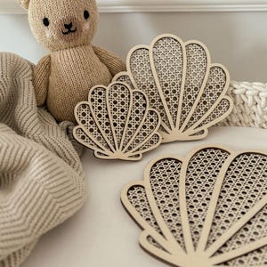 May include: A beige knitted teddy bear sits near three wooden seashell-shaped coasters with a woven cane design. The coasters are arranged on a white surface, with a beige blanket and a white woven basket in the background.