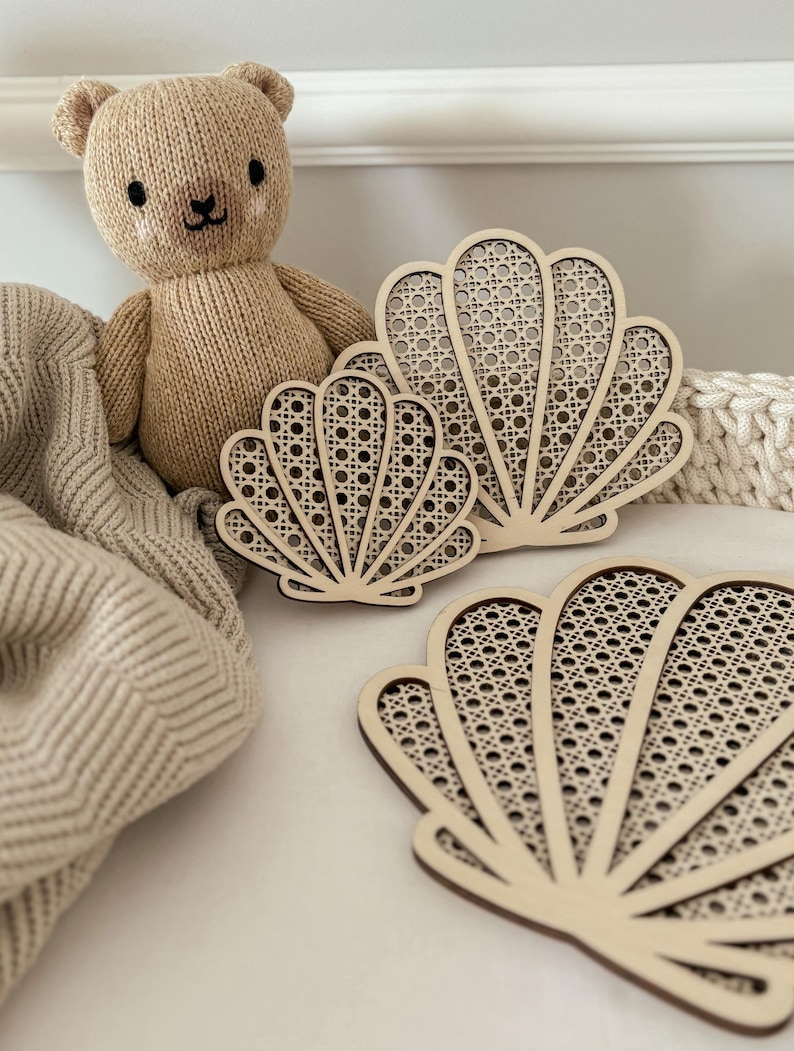 May include: A beige knitted teddy bear sits near three wooden seashell-shaped coasters with a woven cane design. The coasters are arranged on a white surface, with a beige blanket and a white woven basket in the background.