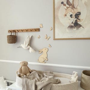 May include: A nursery scene with a white dresser, a woven changing basket, and a beige blanket. A framed illustration of a bunny with a crown hangs on the wall, along with wooden butterflies and a bunny cutout. A teddy bear and a small stuffed animal are in the basket.