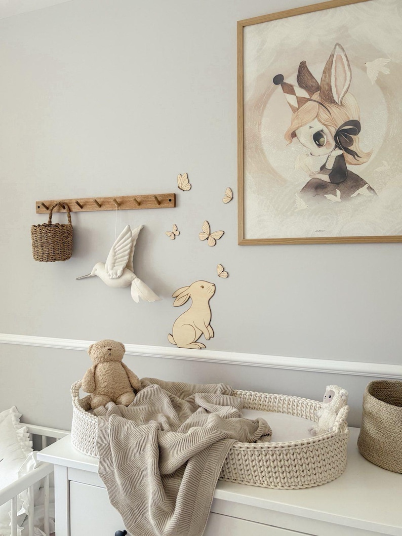 May include: A nursery scene with a white dresser, a woven changing basket, and a beige blanket. A framed illustration of a bunny with a crown hangs on the wall, along with wooden butterflies and a bunny cutout. A teddy bear and a small stuffed animal are in the basket.