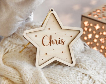 Puede incluir: Un adorno de madera de color claro con forma de estrella, borde recortado y cordón dorado para colgar. El centro tiene grabado el nombre Chris en letra cursiva, rodeado de pequeños detalles de estrellas y puntos grabados en la superficie.