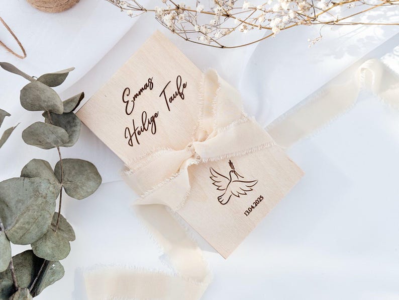 May include: A wooden box with a white ribbon tied around it. The box is engraved with the text "Emma's Holy Taufe" and the date "13.04.2023".