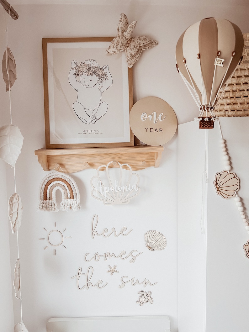 May include: A nursery room decorated with a white and brown hot air balloon, a wooden sign that says "one year", a wooden rainbow, a wooden sun, and a wooden sign that says "here comes the sun".