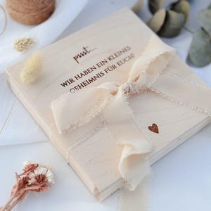 May include: A light-colored wooden box tied with a cream-colored ribbon. The box has text in German and a small heart-shaped design. Dried flowers and twine are visible in the background.