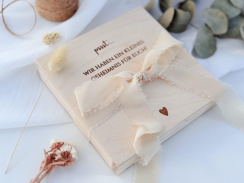 May include: A light-colored wooden box tied with a cream-colored ribbon. The box has text in German and a small heart-shaped design. Dried flowers and twine are visible in the background.