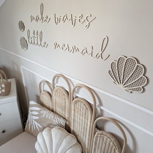 May include: A bedroom scene featuring a bed with a woven headboard and seashell-shaped pillows. The wall above the bed has wooden cutouts that spell out "make waves little mermaid" and seashell accents. The color palette is neutral with beige and white tones.