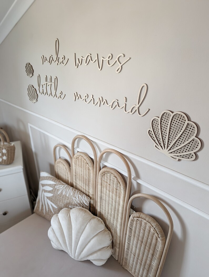 May include: A bedroom scene featuring a bed with a woven headboard and seashell-shaped pillows. The wall above the bed has wooden cutouts that spell out "make waves little mermaid" and seashell accents. The color palette is neutral with beige and white tones.