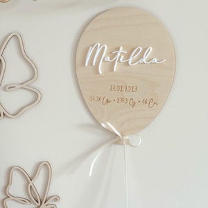May include: A wooden balloon-shaped sign with the name "Matilda" in white script. The sign includes the date 24.02.2023, time, weight, and length. A white ribbon is tied at the bottom. Two wooden butterfly cutouts are on the left.
