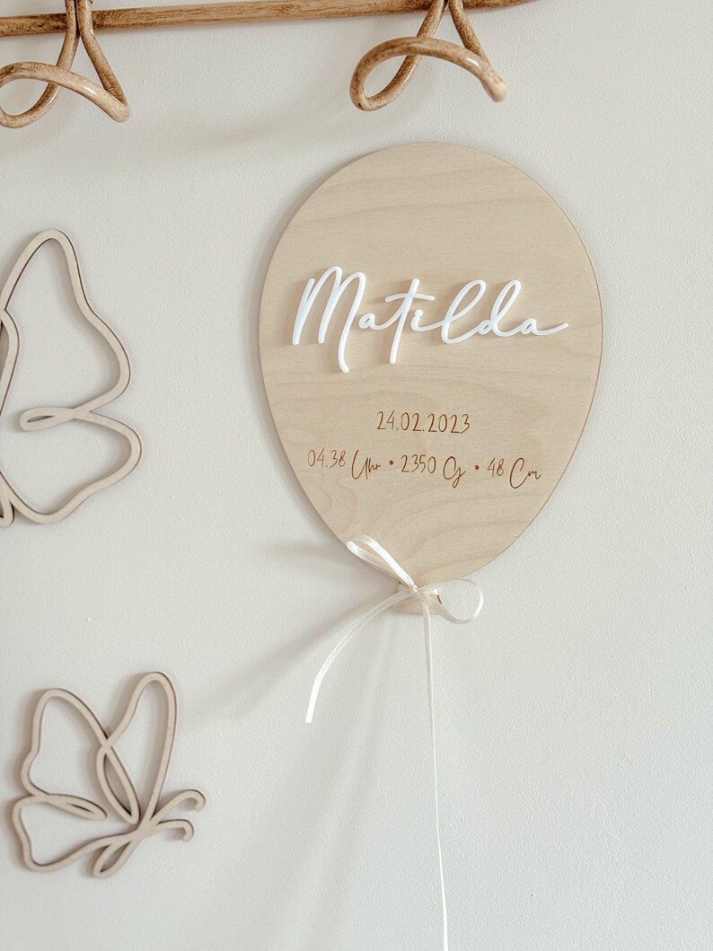 May include: A wooden balloon-shaped sign with the name "Matilda" in white script. The sign includes the date 24.02.2023, time, weight, and length. A white ribbon is tied at the bottom. Two wooden butterfly cutouts are on the left.