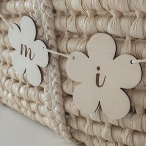 May include: Close-up of a woven basket with a braided handle and a string of wooden flower cutouts. The flower cutouts have the letters "m" and "i" on them. The basket is a light beige color.