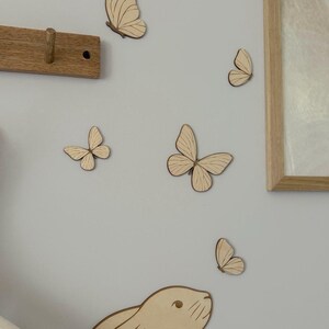 May include: Wooden butterfly and bunny wall decorations in various sizes are arranged on a light gray wall. A wooden coat rack and a framed picture are also visible. The butterflies and bunny are made of light-colored wood.