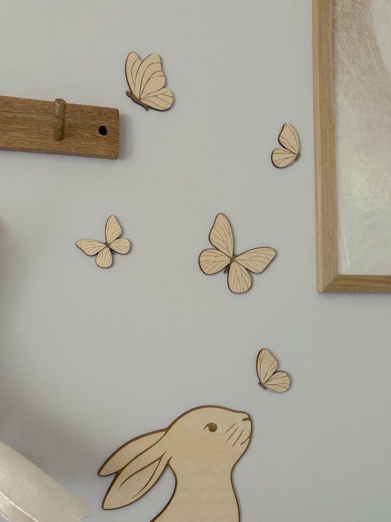 May include: Wooden butterfly and bunny wall decorations in various sizes are arranged on a light gray wall. A wooden coat rack and a framed picture are also visible. The butterflies and bunny are made of light-colored wood.