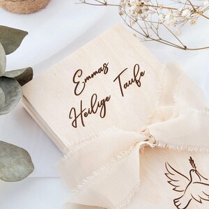 May include: A wooden box with a white ribbon and the text "Emma's Heilige Taufe" engraved on the top.