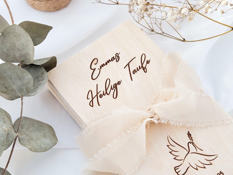 May include: A wooden box with a white ribbon and the text "Emma's Heilige Taufe" engraved on the top.