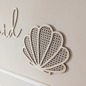 May include: A wooden seashell wall decor piece with a woven cane design. The light-colored wood is cut into a seashell shape and mounted on a neutral-colored wall. The design is a decorative element for a coastal or beach-themed interior.