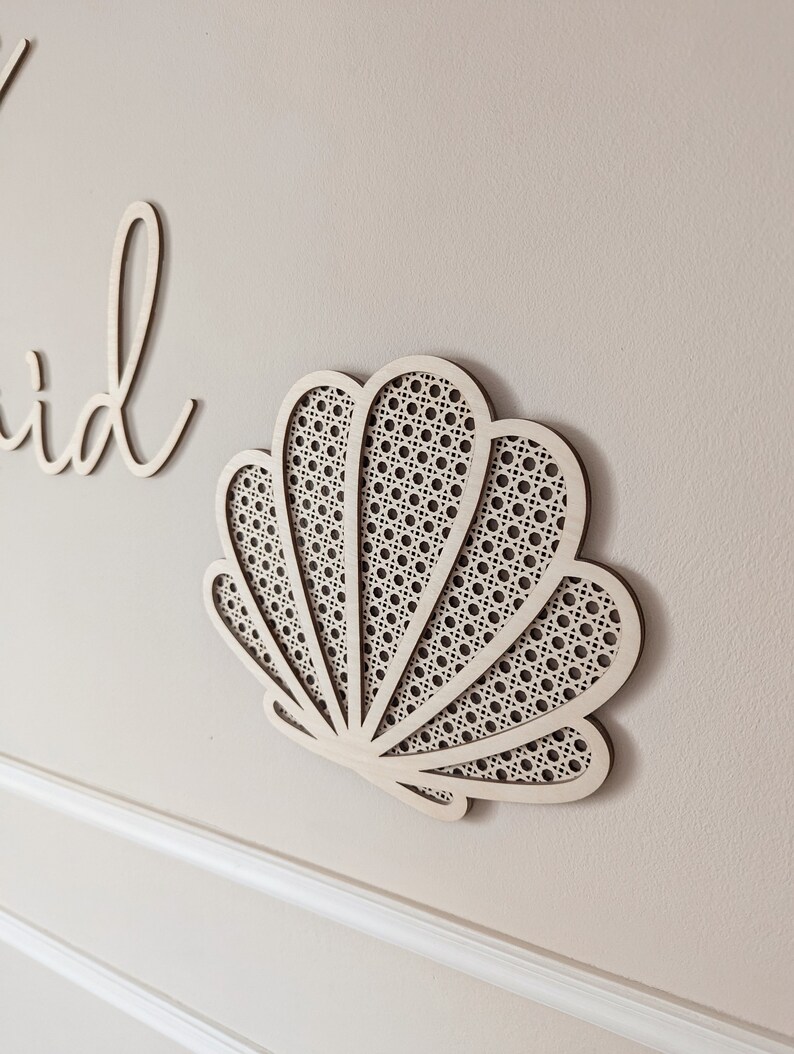 May include: A wooden seashell wall decor piece with a woven cane design. The light-colored wood is cut into a seashell shape and mounted on a neutral-colored wall. The design is a decorative element for a coastal or beach-themed interior.