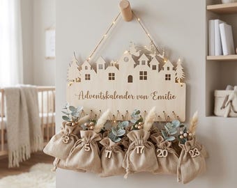 May include: A light-colored wooden advent calendar wall hanging featuring a laser-cut village silhouette, string lights, and the text "Adventskalender von Emilie". Six beige fabric drawstring pouches with wooden numbers and dried foliage hang below.