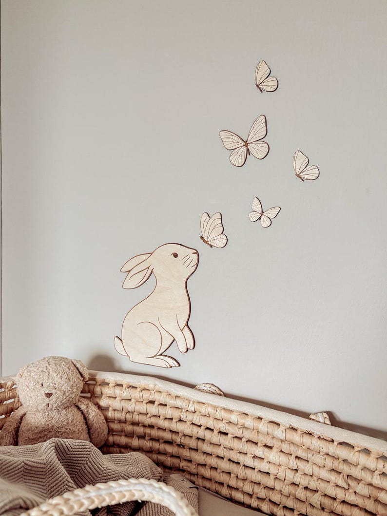 May include: Wooden bunny and butterfly wall decor. The bunny is light brown and appears to be looking up at the butterflies. The butterflies are also light brown and are of varying sizes. A woven basket and teddy bear are in the foreground.