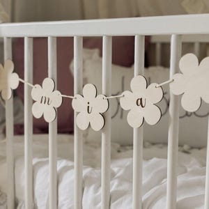May include: A wooden flower garland with the letters "m i a" strung across a white crib. The flowers are light-colored and the letters are in a simple font. The crib is white with white bedding.