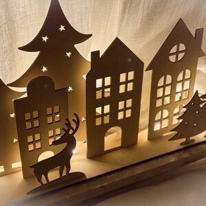 Puede incluir: Escena de pueblo navide&ntilde;o de madera con casas, &aacute;rboles y una silueta de reno iluminados. Las casas tienen ventanas cuadradas y arqueadas, y los &aacute;rboles tienen recortes de estrellas. La escena est&aacute; iluminada desde atr&aacute;s, creando un brillo c&aacute;lido.
