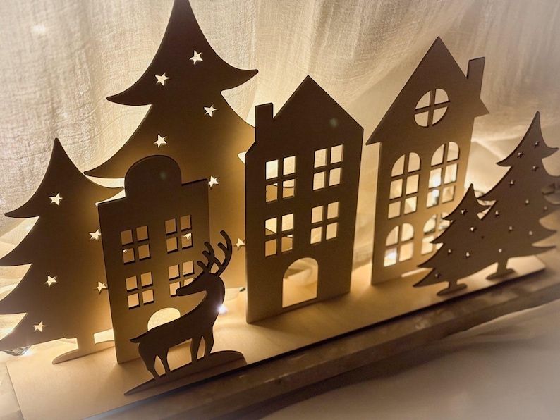 Puede incluir: Escena de pueblo navide&ntilde;o de madera con casas, &aacute;rboles y una silueta de reno iluminados. Las casas tienen ventanas cuadradas y arqueadas, y los &aacute;rboles tienen recortes de estrellas. La escena est&aacute; iluminada desde atr&aacute;s, creando un brillo c&aacute;lido.