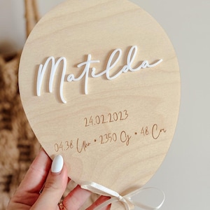 May include: A light wooden balloon-shaped plaque with the name "Matilda" in white script. The date 24.02.2023, time, weight, and height (48 cm) are engraved below. A white ribbon is tied at the bottom.