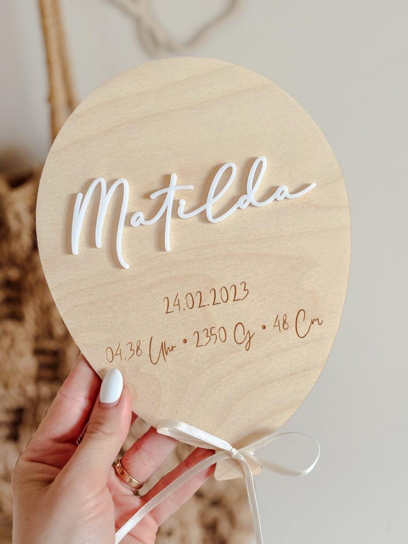 May include: A light wooden balloon-shaped plaque with the name "Matilda" in white script. The date 24.02.2023, time, weight, and height (48 cm) are engraved below. A white ribbon is tied at the bottom.