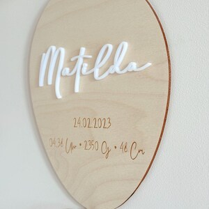 May include: A wooden balloon-shaped sign with the name "Matilda" in white cursive lettering. The sign includes the date 02/24/2023, time, weight, and length (48 cm) details. A white ribbon is tied at the bottom.