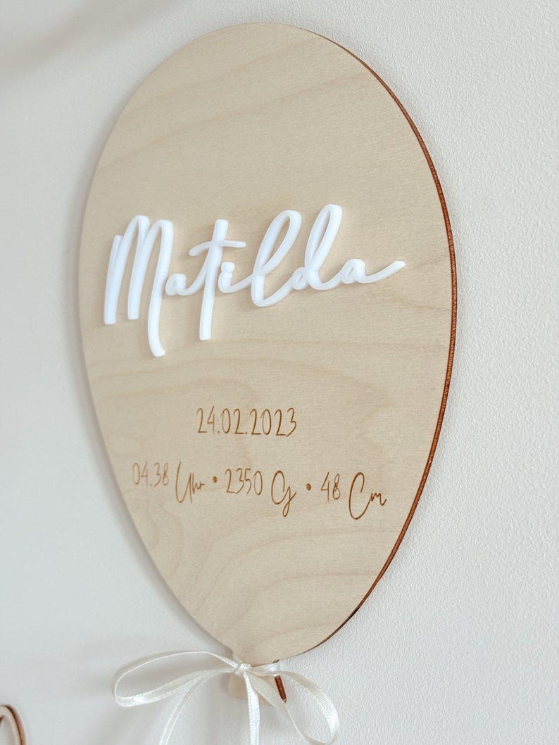 May include: A wooden balloon-shaped sign with the name "Matilda" in white cursive lettering. The sign includes the date 02/24/2023, time, weight, and length (48 cm) details. A white ribbon is tied at the bottom.