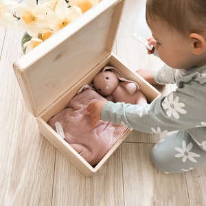 Peut inclure: Une bo&icirc;te en bois avec un pull en tricot rose clair et un jouet de lapin rose &agrave; l'int&eacute;rieur. La bo&icirc;te est ouverte et un enfant met la main dans la bo&icirc;te.